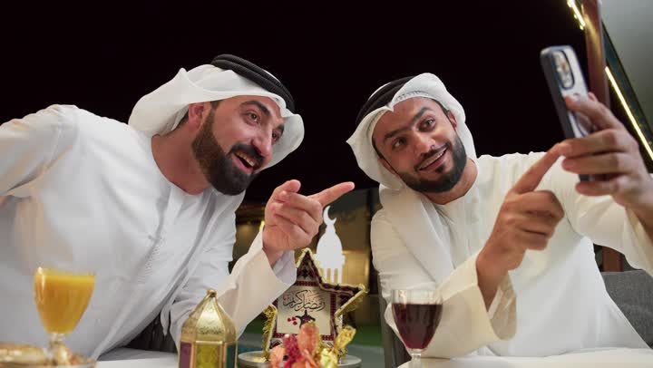 استخدام الهاتف المحمول بتعابير السرور والسعادة، زينة وديكور رمضاني، جلسة ليلية خارجية في احدى شاليهات الامارات، حسن الاستقبال وكرم الضيافة في شهر رمضان المبارك، رجلان عربيان خليجيان اماراتيان يرتديان الكندورة والغترة يجلسان في الهواء الطلق، تصفح مواقع الانترنت