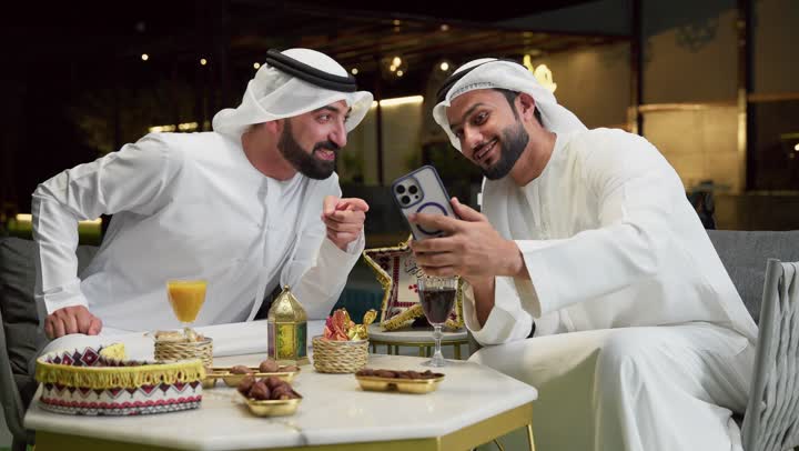 قضاء وقت ممتع مع الأصدقاء، استخدام الهاتف المحمول بتعابير السرور والسعادة، زينة وديكور رمضاني، حسن الاستقبال وكرم الضيافة في شهر رمضان المبارك، رجلان عربيان خليجيان اماراتيان يرتديان الكندورة والغترة يجلسان في شاليه سياحي، تصفح مواقع الانترنت