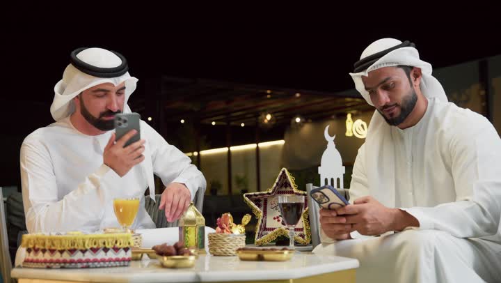 إجراء مكالمة هاتفية، حسن الاستقبال وكرم الضيافة في شهر رمضان المبارك، زينة وديكور رمضاني، جلسة ليلية خارجية في احدى شاليهات الامارات، صديقان عربيان خليجيان اماراتيان يرتديان الكندورة والغترة يجلسان في الهواء الطلق ويستخدمان الهاتف المحمول بتعابير الاندماج