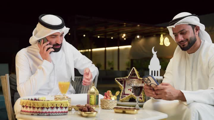 الرد على مكالمة هاتفية، حسن الاستقبال وكرم الضيافة في شهر رمضان المبارك، زينة وديكور رمضاني، جلسة ليلية خارجية في احدى شاليهات الامارات، صديقان عربيان خليجيان اماراتيان يرتديان الكندورة والغترة يجلسان في الهواء الطلق ويستخدمان الهاتف المحمول بتعابير الاندماج