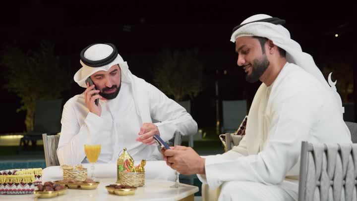 إجراء مكالمة هاتفية، حسن الاستقبال وكرم الضيافة في شهر رمضان المبارك، زينة وديكور رمضاني، جلسة ليلية خارجية في احدى شاليهات الامارات، صديقان عربيان خليجيان اماراتيان يرتديان الكندورة والغترة يجلسان في الهواء الطلق ويستخدمان الهاتف المحمول بتعابير الاندماج