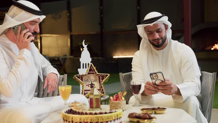 الرد على مكالمة هاتفية، حسن الاستقبال وكرم الضيافة في شهر رمضان المبارك، زينة وديكور رمضاني، جلسة ليلية خارجية في احدى شاليهات الامارات، صديقان عربيان خليجيان اماراتيان يرتديان الكندورة والغترة يجلسان في الهواء الطلق ويستخدمان الهاتف المحمول بتعابير الاندماج