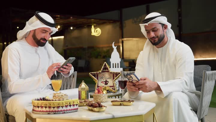 حسن الاستقبال وكرم الضيافة في شهر رمضان المبارك، تصفح مواقع الانترنت، زينة وديكور رمضاني، جلسة ليلية خارجية في احدى شاليهات الامارات، صديقان عربيان خليجيان اماراتيان يرتديان الكندورة والغترة يجلسان في الهواء الطلق ويستخدمان الهاتف المحمول بتعابير الاندماج