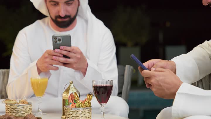 تصفح مواقع الانترنت، زينة واضاءة وديكور رمضاني، جلسة ليلية خارجية في احدى شاليهات الامارات، حسن الاستقبال وكرم الضيافة في شهر رمضان المبارك، صديقان عربيان خليجيان اماراتيان يرتديان الكندورة والغترة يجلسان في الهواء الطلق ويستخدمان الهاتف المحمول بتعابير الاندماج