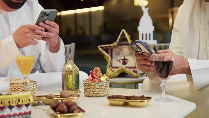 تصفح مواقع الانترنت، زينة واضاءة وديكور رمضاني، جلسة ليلية خارجية في احدى شاليهات الامارات، حسن الاستقبال وكرم الضيافة في شهر رمضان المبارك، صديقان عربيان خليجيان اماراتيان يرتديان الكندورة والغترة يجلسان في الهواء الطلق ويستخدمان الهاتف المحمول بتعابير الاندماج