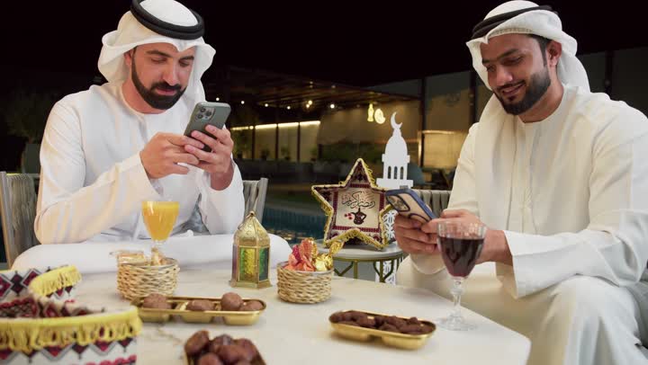 Warm welcome and hospitality in the holy month of Ramadan, browsing the internet, Ramadan decorations, an outdoor night session in one of the chalets in the Emirates, two Emirati Gulf Arab friends wearing kanduras and ghutras sitting outdoors and using mobile phones with expressions of integration