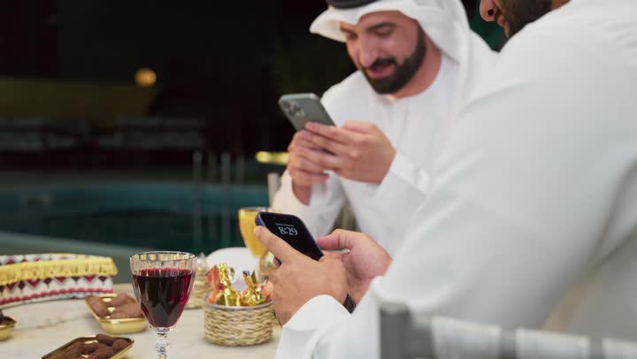 حسن الاستقبال وكرم الضيافة في شهر رمضان المبارك، تصفح مواقع الانترنت، زينة وديكور رمضاني، جلسة ليلية خارجية في احدى شاليهات الامارات، صديقان عربيان خليجيان اماراتيان يرتديان الكندورة والغترة يجلسان في الهواء الطلق ويستخدمان الهاتف المحمول بتعابير الاندماج