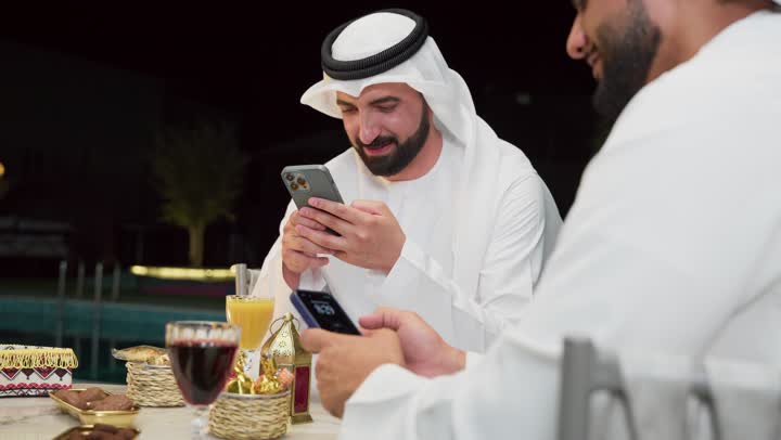 Warm welcome and hospitality in the holy month of Ramadan, browsing the internet, Ramadan decorations, an outdoor night session in one of the chalets in the Emirates, two Emirati Gulf Arab friends wearing kanduras and ghutras sitting outdoors and using mobile phones with expressions of integration