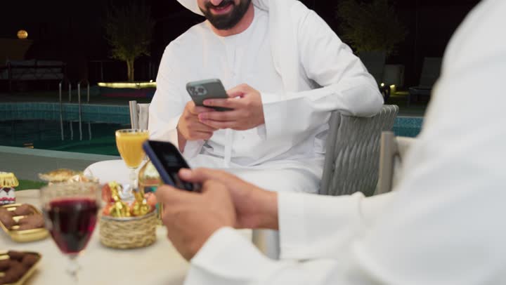 تصفح مواقع الانترنت، زينة واضاءة وديكور رمضاني، جلسة ليلية خارجية في احدى شاليهات الامارات، حسن الاستقبال وكرم الضيافة في شهر رمضان المبارك، صديقان عربيان خليجيان اماراتيان يرتديان الكندورة والغترة يجلسان في الهواء الطلق ويستخدمان الهاتف المحمول بتعابير الاندماج