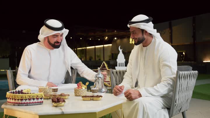 جلسة ليلية خارجية في إحدى شاليهات الامارات، حسن الاستقبال وكرم الضيافة في شهر رمضان، صديقان عربيان خليجيان اماراتيان يرتديان الكندورة والغترة يجلسان في الهواء الطلق يتبادلان الاحاديث والاخبار بتعابير السرور، رجل سعودي يقوم بصب القهوة العربية من الدلة الفضية