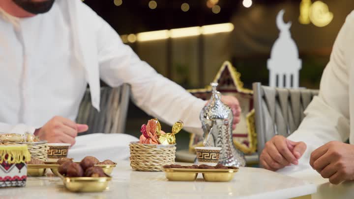 Warm welcome and hospitality in the month of Ramadan, an outdoor night gathering in one of the chalets in the Emirates, two Emirati Gulf Arab friends wearing kanduras and ghutras sitting in the open air exchanging conversations and news with expressions of pleasure, a close-up of a Saudi man pouring Arabic coffee