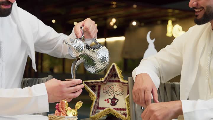 Warm welcome and hospitality in the month of Ramadan, an outdoor night gathering in one of the chalets in the Emirates, two Emirati Gulf Arab friends wearing kanduras and ghutras sitting in the open air exchanging conversations and news with expressions of pleasure, a close-up of a Saudi man pouring Arabic coffee