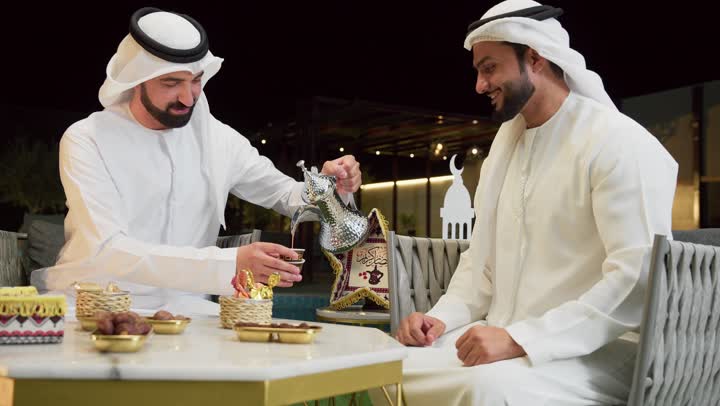 جلسة ليلية خارجية في إحدى شاليهات الامارات، حسن الاستقبال وكرم الضيافة في شهر رمضان، صديقان عربيان خليجيان اماراتيان يرتديان الكندورة والغترة يجلسان في الهواء الطلق يتبادلان الاحاديث والاخبار بتعابير السرور، رجل سعودي يقوم بصب القهوة العربية من الدلة الفضية