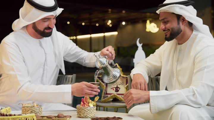 جلسة ليلية خارجية في إحدى شاليهات الامارات، حسن الاستقبال وكرم الضيافة في شهر رمضان، صديقان عربيان خليجيان اماراتيان يرتديان الكندورة والغترة يجلسان في الهواء الطلق يتبادلان الاحاديث والاخبار بتعابير السرور، رجل سعودي يقوم بصب القهوة العربية من الدلة الفضية