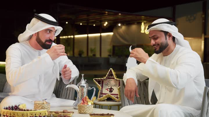 Ramadan decor and accessories, an outdoor night session in one of the chalets in the Emirates, warm welcome and hospitality in the holy month of Ramadan, two Emirati Gulf Arab friends wearing kanduras and ghutras sitting outdoors drinking Arabic coffee, exchanging conversations and news with expressions of pleasure