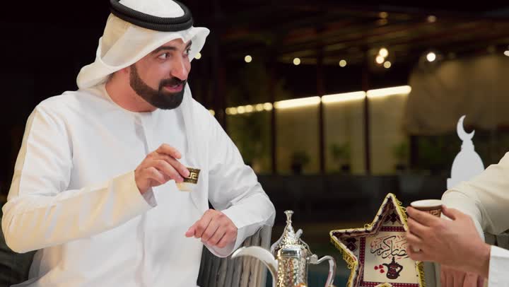 ديكور واكسسوات رمضانية، جلسة ليلية خارجية في إحدى شاليهات الامارات، حسن الاستقبال وكرم الضيافة في شهر رمضان المبارك، صديقان عربيان خليجيان اماراتيان يرتديان الكندورة والغترة يجلسان في الهواء الطلق ويتناولان القهوة العربية، تبادل الاحاديث والاخبار بتعابير السرور
