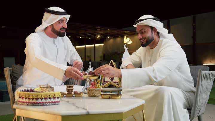 Spending a pleasant time with friends, warm welcome and hospitality in the month of Ramadan, an outdoor night gathering in one of the chalets in the Emirates, two Arab Gulf Emirati friends wearing kanduras and ghutras sitting in the open air exchanging conversations and news with expressions of pleasure, eating dates to break the fast