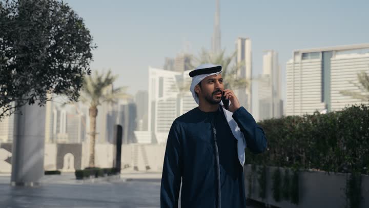 Integrating modern technologies into daily life, chatting and communicating with family, making a phone call via mobile phone, an Emirati Gulf Arab man spending a pleasant time in a park in the Emirates, a tour in nature, an outdoor picnic, hearing news and happy surprises