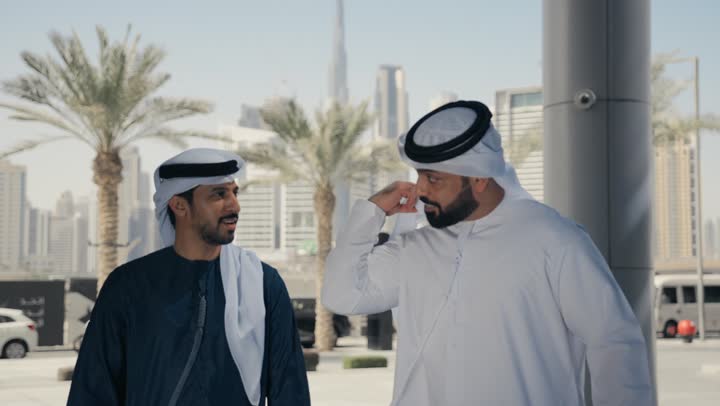 الاستمتاع والتجول مع الاصدقاء ، قضاء اوقات ممتعه ، مفهوم الأناقة والاهتمام بالمظهر الخارجي ، المشي فى الخارج ، استنشاق الهواء النقي في وقت النهار، صديقان عربيان خليجيان اماراتيان يرتديان الكندوره والغتره ، النظر الى بعضهما بتعابير السرور.