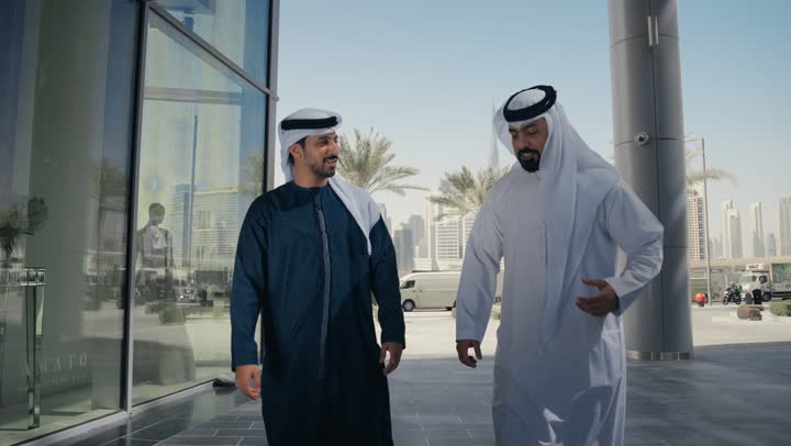 Dialogue and discussion between friends, the concept of elegance and interest in one's outward appearance, walking outside, enjoying, strolling and breathing fresh air during the day, two Arab Gulf Emirati friends wearing kanduras and ghutras, looking at each other with expressions of pleasure.