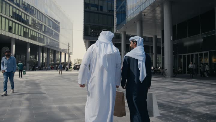 الوقوف بثقة في شوارع المدينة، مفهوم الأناقة والاهتمام بالمظهر الخارجي ، المشي فى الخارج ،الاستمتاع والتجول واستنشاق الهواء النقي في وقت النهار، صديقان عربيان خليجيان اماراتيان يرتديان الكندوره والغتره ، النظر الى بعضهما بتعابير السرور.