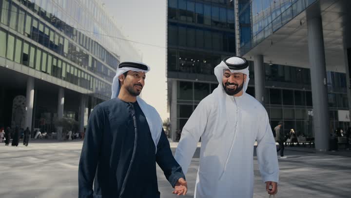 The concept of elegance and attention to one's appearance, walking outside, standing confidently in the city streets, enjoying, strolling and breathing fresh air during the day, two Arab Gulf Emirati friends wearing kandoras and ghutras, looking at each other with expressions of pleasure.