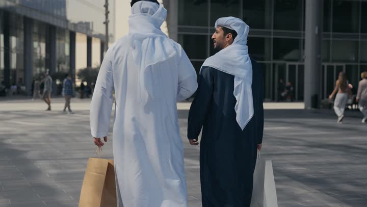 المشي فى الخارج ، مفهوم الأناقة والاهتمام بالمظهر الخارجي ، الوقوف بثقة في شوارع المدينة، الاستمتاع والتجول واستنشاق الهواء النقي في وقت النهار، صديقان عربيان خليجيان اماراتيان يرتديان الكندوره والغتره ، النظر الى بعضهما بتعابير السرور.