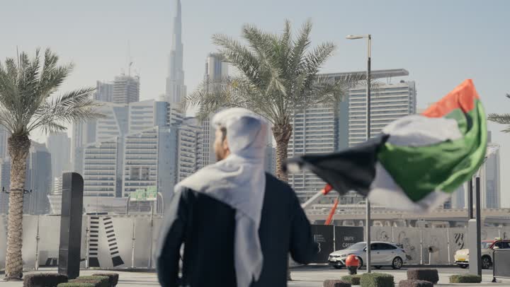 Commemorating the country's independence, gestures of pride and happiness, love of the homeland and belonging to it, a picture of a man wearing a kandura and ghutra raising the flag of the United Arab Emirates, UAE Independence Day, learning about the cultures and history of the people of the Emirates, celebrating National Day, December 2