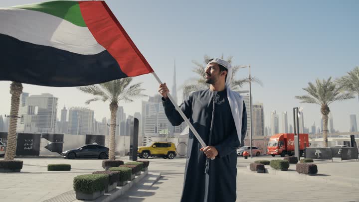 Gestures of pride and happiness, love of the homeland and belonging to it, a picture of a man wearing a kandura and ghutra raising the flag of the United Arab Emirates, commemorating the country's independence, UAE Independence Day, learning about the cultures and history of the people of the Emirates, celebrating National Day, December 2