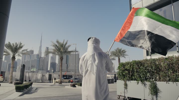 Learning about the culture and history of the Emirati people, commemorating the country's independence, gestures of pride and happiness, love of the homeland and belonging to it, a picture of a man wearing a kandura and ghutra raising the flag of the United Arab Emirates, UAE Independence Day, celebrating National Day, December 2