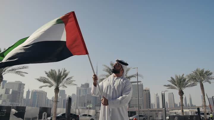 A picture of a man wearing a kandura and ghutra raising the flag of the United Arab Emirates, UAE Independence Day, National Day celebrations December 2, gestures of pride and happiness, the anniversary of the founding of the Union 1971, learning about the cultures and history of the people of the Emirates, reviving the love of the homeland and belonging to it