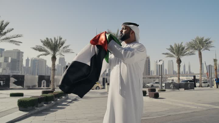 Learning about the cultures and history of the people of the Emirates, UAE Independence Day, love of the homeland and belonging to it, National Day December 2, a picture of a man wearing a kandura and ghutra, placing the flag of the United Arab Emirates on his shoulder
