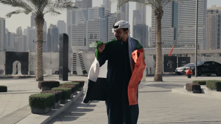 Standing up straight and looking at the camera with proud gestures, UAE Independence Day, love and belonging to the homeland, celebrating the National Day on December 2, learning about the culture and history of the people of the Emirates, a photo of a man wearing a kandura and ghutra with the flag of the United Arab Emirates on his shoulder