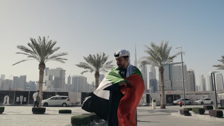 يوم استقلال الإمارات ، حب الوطن والانتماء إليه, الاحتفال باليوم الوطني 2 ديسمبر،التعرف على ثقافات وتاريخ شعب الامارات، تصوير لرجل يرتدي الكندوره والغتره يضع علم دولة الامارات العربية المتحدة على كتفه، الوقوف باستقامة والنظر إلى الكاميرا بإيماءات الفخر
