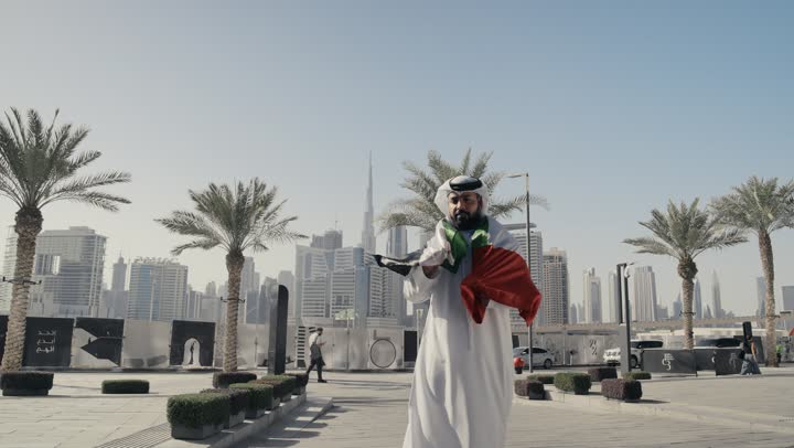 هوية اليوم الوطني الإماراتي 51 ، حب الوطن والانتماء إليه, الاحتفال باليوم الوطني 2 ديسمبر،التعرف على ثقافات وتاريخ شعب الامارات، تصوير لرجل يرتدي الكندوره والغتره يضع علم دولة الامارات العربية المتحدة على كتفه، الوقوف باستقامة والنظر إلى الكاميرا بإيماءات الفخر