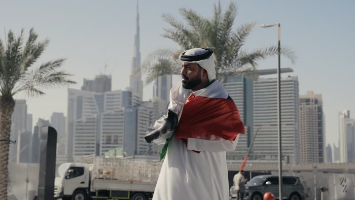 Showing feelings of pride in the Emirati identity, love of the homeland and belonging to it, celebrating the National Day on December 2, learning about the cultures and history of the people of the Emirates, photographing a man wearing a kandura and ghutra, placing the flag of the United Arab Emirates on his shoulder, standing up straight and looking at the camera with gestures of pride