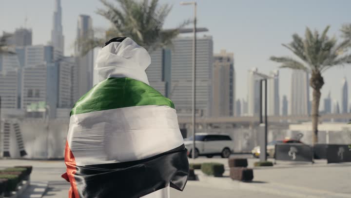 حب الوطن والانتماء إليه, إظهار مشاعر الفخر بالهوية الاماراتية، الاحتفال باليوم الوطني 2 ديسمبر،التعرف على ثقافات وتاريخ شعب الامارات، تصوير لرجل يرتدي الكندوره والغتره يضع علم دولة الامارات العربية المتحدة على كتفه، الوقوف باستقامة والنظر إلى الكاميرا بإيماءات الفخر