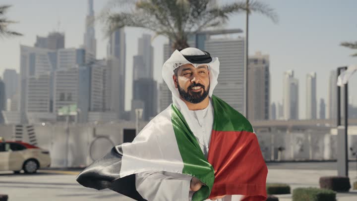 إحياء ذكرى استقلال البلاد، الاحتفال باليوم الوطني 2 ديسمبر،التعرف على ثقافات وتاريخ شعب الامارات،  تصوير لرجل يرتدي الكندوره والغتره يضع علم دولة الامارات العربية المتحدة على كتفه، الوقوف باستقامة والنظر إلى الكاميرا بإيماءات الفخر