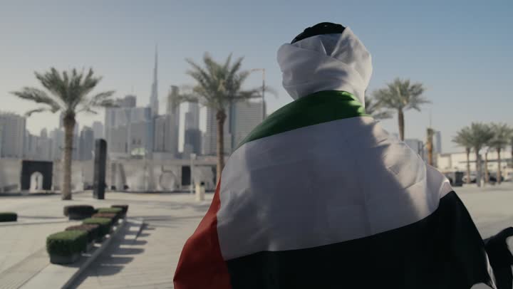 الاحتفال باليوم الوطني 2 ديسمبر،التعرف على ثقافات وتاريخ شعب الامارات، إحياء ذكرى استقلال البلاد، تصوير لرجل يرتدي الكندوره والغتره يضع علم دولة الامارات العربية المتحدة على كتفه، الوقوف باستقامة والنظر إلى الكاميرا بإيماءات الفخر