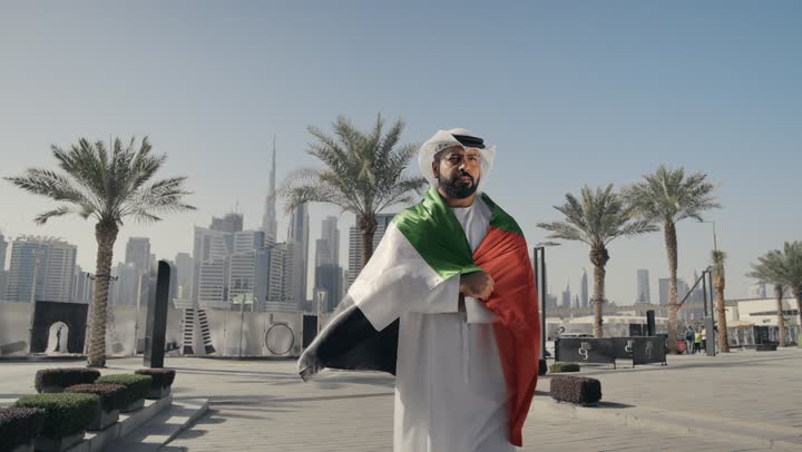 التعرف على ثقافات وتاريخ شعب الامارات، الاحتفال باليوم الوطني 2 ديسمبر، إحياء ذكرى استقلال البلاد، تصوير لرجل يرتدي الكندوره والغتره يضع علم دولة الامارات العربية المتحدة على كتفه، الوقوف باستقامة والنظر إلى الكاميرا بإيماءات الفخر