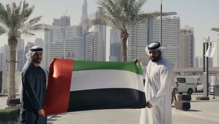 Celebrating National Day, December 2, the UAE's Independence Day, learning about the culture and history of the Emirati people, love of the homeland and belonging to it, a photo of two men wearing kandoras and ghutras holding the flag of the United Arab Emirates