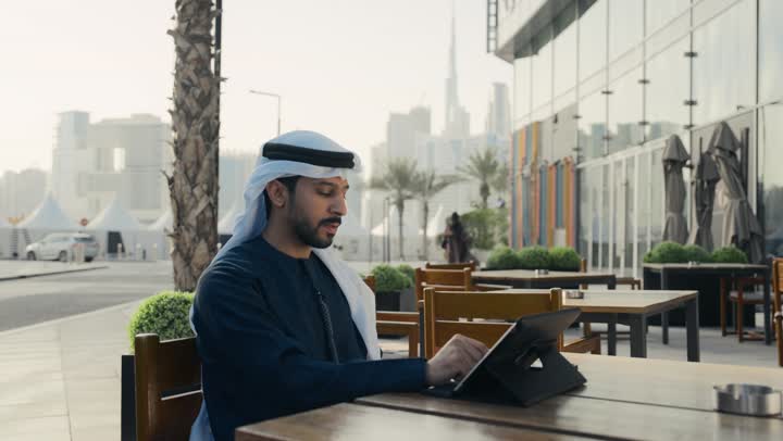 قضاء اوقات ممتعه ، تصفح مواقع التواصل الاجتماعي عن طريق الانترنت ، الاستمتاع بالاجواء الهادئة خارج المنزل، مطاعم و كافيهات في الإمارات العربية المتحدة، رجل عربي خليجي اماراتي يرتدي الكندورة والغترة البيضاء يجلس على كرسي في إحدى المقاهي الراقية يقوم بتصفح التابلت