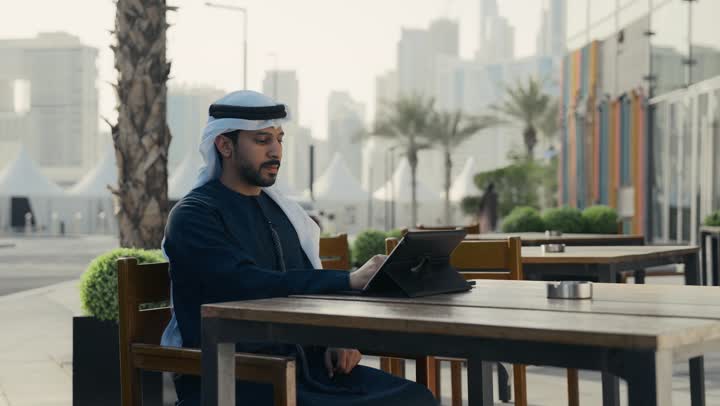 تصفح تطبيقات وبرامج التابلت ، تصفح مواقع التواصل الاجتماعي عن طريق الانترنت ، الاستمتاع بالاجواء الهادئة خارج المنزل، مطاعم و كافيهات في الإمارات العربية المتحدة،رجل عربي خليجي اماراتي يرتدي الكندورة والغترة البيضاء يجلس على كرسي في إحدى المقاهي الراقية يقوم بتصفح التابلت