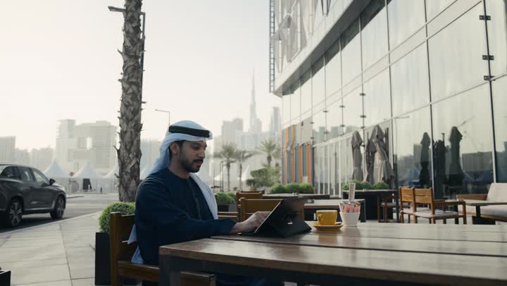 انجاز المهام عن بعد ، تصفح مواقع التواصل الاجتماعي عن طريق الانترنت ، الاستمتاع بالاجواء الهادئة خارج المنزل، مطاعم و كافيهات في الإمارات العربية المتحدة،رجل عربي خليجي اماراتي يرتدي الكندورة والغترة البيضاء يجلس على كرسي في إحدى المقاهي الراقية يقوم بتصفح التابلت
