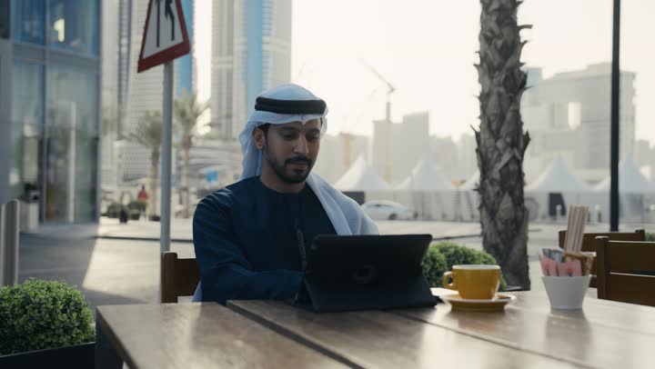 تصفح مواقع التواصل الاجتماعي عن طريق الانترنت ، الاستمتاع بالاجواء الهادئة خارج المنزل، مطاعم و كافيهات في الإمارات العربية المتحدة،رجل عربي خليجي اماراتي يرتدي الكندورة والغترة البيضاء يجلس على كرسي في إحدى المقاهي الراقية يقوم بتصفح التابلت