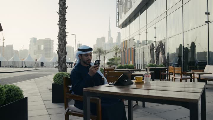 الاستمتاع بالاجواء الهادئة خارج المنزل، مطاعم و كافيهات في الإمارات العربية المتحدة،رجل عربي خليجي اماراتي يرتدي الكندورة والغترة البيضاء يجلس على كرسي في إحدى المقاهي الراقية يقوم بتصفح التابلت ، تصفح مواقع التواصل الاجتماعي عن طريق الانترنت