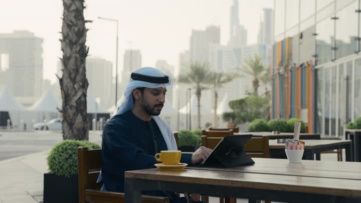 مطاعم و كافيهات في الإمارات العربية المتحدة، الاستمتاع بالاجواء الهادئة خارج المنزل، رجل عربي خليجي اماراتي يرتدي الكندورة والغترة البيضاء يجلس على كرسي في إحدى المقاهي الراقية يقوم بتصفح التابلت ، تصفح مواقع التواصل الاجتماعي عن طريق الانترنت
