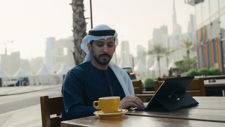 مطاعم وكافيهات فى الامارات، إنجاز مهام العمل عن بعد، إدارة شؤون الأعمال وتنظيمها ، تصوير لرجل عربي خليجي اماراتي يرتدي الكندورة والغترة يجلس على كرسي في إحدى المقاهي يشرب مشروب ساخن ويستخدم الحاسوب المحمول ، الاستمتاع بشرب القهوه ، استخدام التكنولوجيا والتقنيات المتطورة .
