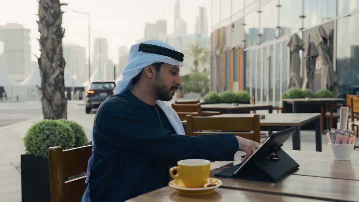 إنجاز مهام العمل عن بعد، إدارة شؤون الأعمال وتنظيمها ، تصوير لرجل عربي خليجي اماراتي يرتدي الكندورة والغترة يجلس على كرسي في إحدى المقاهي يشرب مشروب ساخن ويستخدم الحاسوب المحمول ، الاستمتاع بشرب القهوه ، استخدام التكنولوجيا والتقنيات المتطورة .
