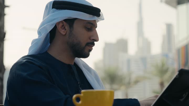 إدارة شؤون الأعمال وتنظيمها ، تصوير لرجل عربي خليجي اماراتي يرتدي الكندورة والغترة يجلس على كرسي في إحدى المقاهي يشرب مشروب ساخن ويستخدم الحاسوب المحمول ، الاستمتاع بشرب القهوه ، إنجاز مهام العمل عن بعد، استخدام التكنولوجيا والتقنيات المتطورة .